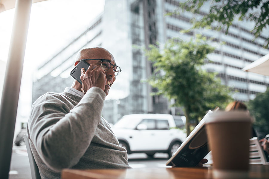 Contact - Portrait of a Smilinig Mature Businessman Sitting in a Cafe While Talking on the Phone with Views of a Commercial Building Through the Window
